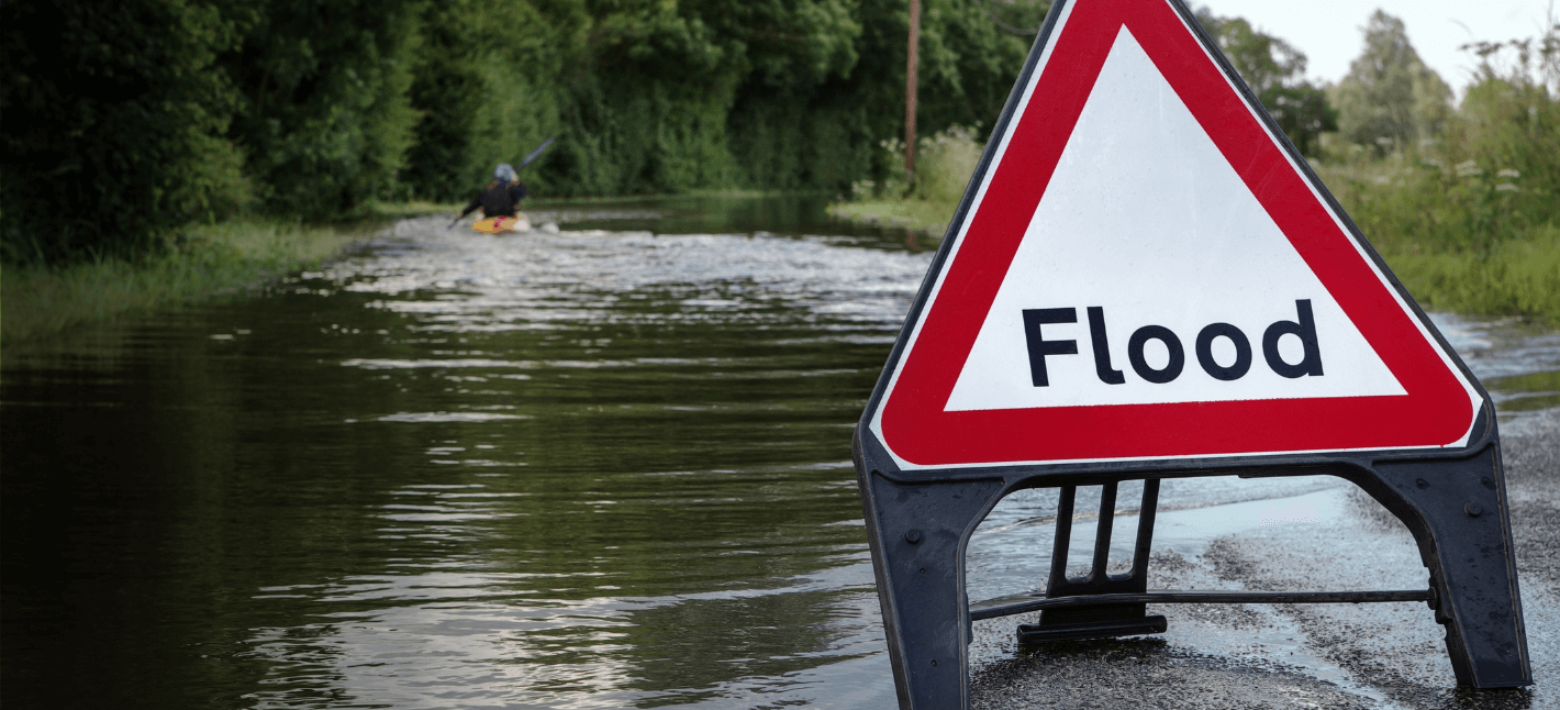 Image shows a warning street sign with a word "flood" and the flooded street behind it